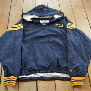 Könnte beinhalten: Marineblaue Kapuzenjacke mit gelben Streifen an Ärmeln und Bündchen. Die Jacke hat das UCLA-Logo in Gelb auf der Brust und ein Champion-Logo unten rechts. Die Jacke hat einen halben Reißverschluss und ein weißes Futter.