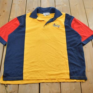 Può includere: Una polo vintage con un design a blocchi di colore. La maglia presenta un pannello centrale giallo, pannelli laterali blu navy e maniche rosse. Il colletto e i polsini sono blu navy. La maglia ha un piccolo logo ricamato sul petto.