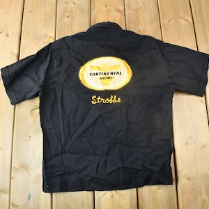 Peut inclure: Une chemise noire à manches courtes avec un écusson brodé en or qui dit "Continental Airlines" et le nom "Strobbe" en dessous.