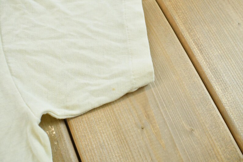 May include: A plain, off-white short-sleeved t-shirt made of a soft, lightweight fabric. The garment features a simple crew neckline and a standard hemmed sleeve, showcasing a clean, solid-colored design with a subtle, light texture throughout.