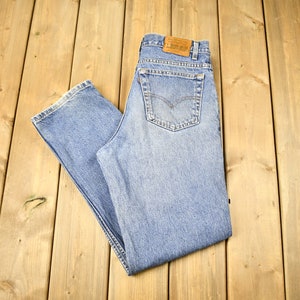 May include: A pair of light blue denim jeans with a brown leather patch on the back waistband. The jeans are folded in half and laid flat on a wooden surface.