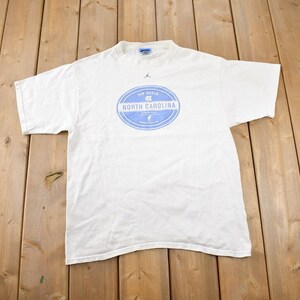 May include: White t-shirt with a blue and white circular logo that says "Tar Heels" and "North Carolina".
