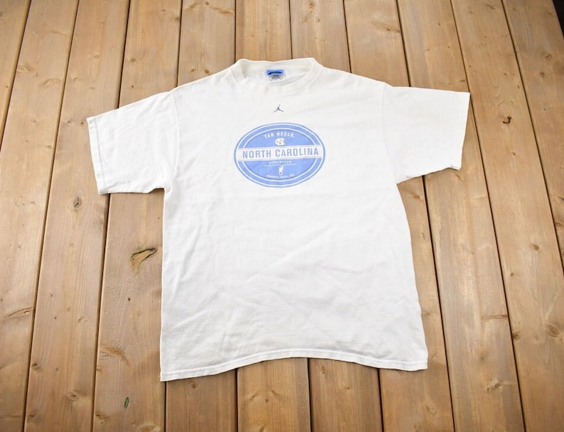 May include: White t-shirt with a blue and white circular logo that says "Tar Heels" and "North Carolina".