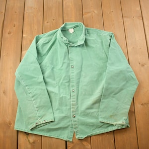 Puede incluir: Una chaqueta de trabajo verde con mangas largas y cierre de botones. La chaqueta tiene un bolsillo en el pecho y está hecha de un tejido resistente.