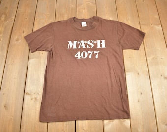 May include: A brown t-shirt with a white print that says "M*A*S*H 4077".