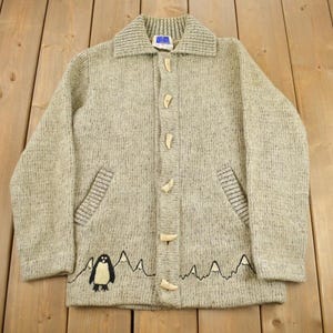 May include: A light gray knit cardigan with a collared neckline and wooden toggle closures. The sweater features two front pockets and a mountain and penguin graphic at the bottom. The cardigan is laid out on a wooden surface.