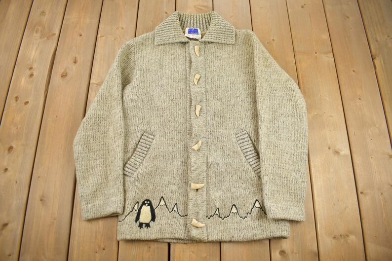 May include: A light gray knit cardigan with a collared neckline and wooden toggle closures. The sweater features two front pockets and a mountain and penguin graphic at the bottom. The cardigan is laid out on a wooden surface.