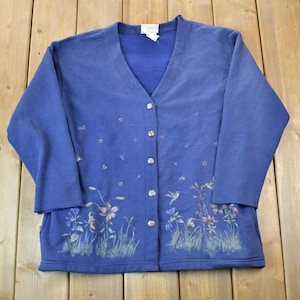 Könnte beinhalten: Ein blauer Cardigan mit einem Blumen- und Kolibri-Design. Der Cardigan hat einen V-Ausschnitt und einen Knopfverschluss.