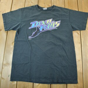 Pode incluir: T-shirt cinza escuro com o texto "Devil Rays" em uma fonte estilizada e um gráfico de arraia. O texto é em azul, roxo e amarelo. A camisa é feita de um material macio.