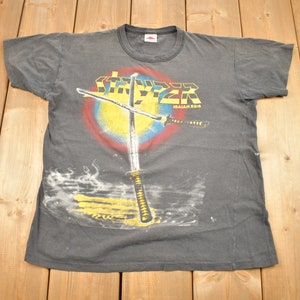 Op de afbeelding: Een zwart T-shirt met een grafische print van twee gekruiste zwaarden boven een gele zon met een rode, blauwe en gele ring eromheen. De tekst "Stryper" is in gele en zwarte letters boven de zwaarden gedrukt en de tekst "Isaiah 53:5" is in zwarte letters onder de zwaarden gedrukt.