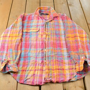 May include: A long-sleeve button-down shirt featuring a vibrant plaid pattern in shades of pink, orange, yellow, and light blue. The shirt includes a standard collar, a single chest pocket on the left side, and buttoned cuffs on the sleeves.