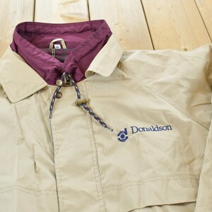 May include: Tan jacket with a maroon collar and a drawstring hood. The jacket has a button closure and the word "Donaldson" embroidered in blue on the front. The jacket is made of a lightweight material.