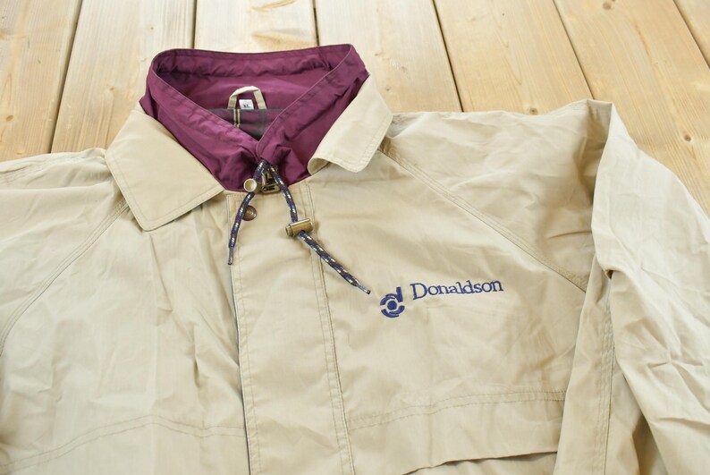 May include: Tan jacket with a maroon collar and a drawstring hood. The jacket has a button closure and the word "Donaldson" embroidered in blue on the front. The jacket is made of a lightweight material.