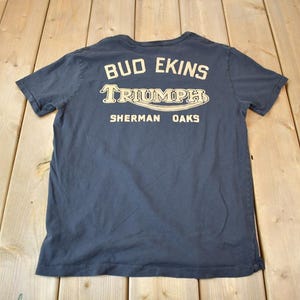 May include: A dark blue t-shirt with a white graphic print that says "Bud Ekins Triumph Sherman Oaks".