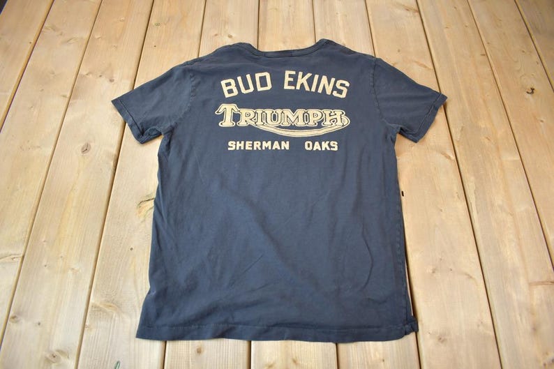 May include: A dark blue t-shirt with a white graphic print that says "Bud Ekins Triumph Sherman Oaks".