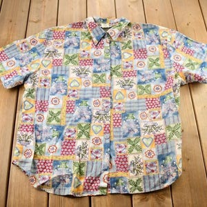 Puede incluir: Una camisa de manga corta con botones azul y blanca con un diseño de patchwork que presenta varios patrones florales y geométricos.