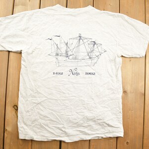 May include: A white t-shirt with a black and white line drawing of a ship on the back. The ship is labeled "Niña" and the years "1492" and "1992" are printed below the ship.