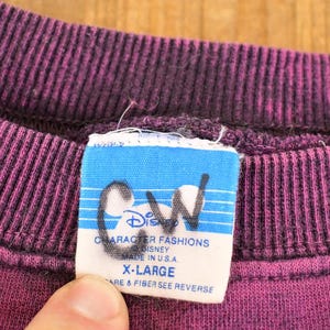 May include: A purple crewneck sweatshirt with a ribbed collar. A white rectangular clothing tag is attached to the inside neckline, featuring the text Disney Character Fashions, Made in U.S.A., and X-Large, with the letters CW written in black ink.