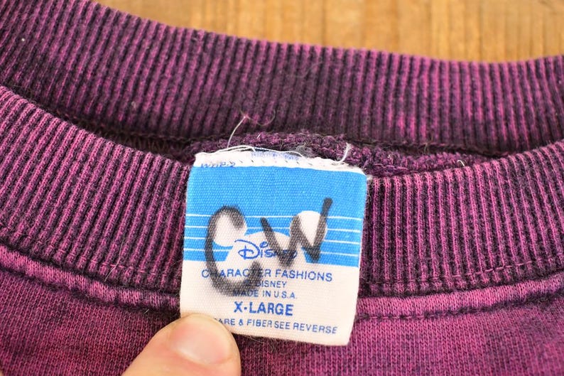 May include: A purple crewneck sweatshirt with a ribbed collar. A white rectangular clothing tag is attached to the inside neckline, featuring the text Disney Character Fashions, Made in U.S.A., and X-Large, with the letters CW written in black ink.