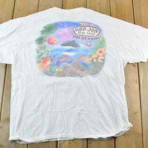 Puede incluir: Camiseta blanca con un gr&aacute;fico colorido de un delf&iacute;n saltando del oc&eacute;ano, rodeado de elementos tropicales. La camiseta tiene el texto "Ron Jon Surf Shop ONE OF A KIND" y "Cocoa Beach, Fla."