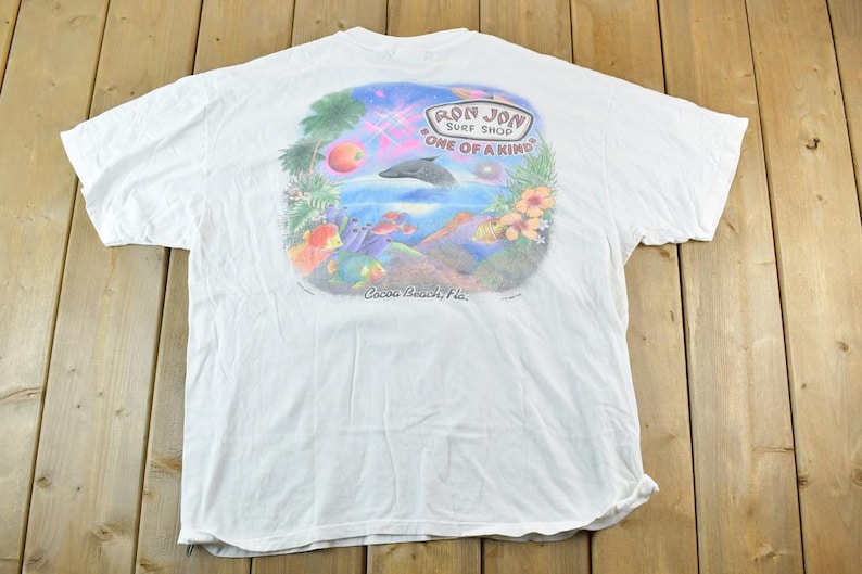 Puede incluir: Camiseta blanca con un gr&aacute;fico colorido de un delf&iacute;n saltando del oc&eacute;ano, rodeado de elementos tropicales. La camiseta tiene el texto "Ron Jon Surf Shop ONE OF A KIND" y "Cocoa Beach, Fla."