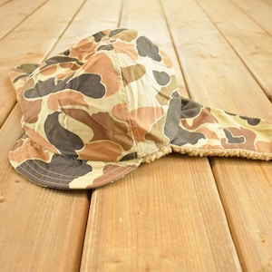 Può includere: Un cappello con motivo mimetico con paraorecchie. Il cappello presenta un motivo marrone, beige e grigio. I paraorecchie sono foderati con un materiale morbido marrone chiaro. Il cappello è progettato per fornire calore e protezione.
