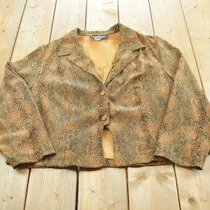 May include: A cropped blazer featuring a brown and black snakeskin pattern, a notched lapel collar, long sleeves, and two front buttons. The garment has a solid tan interior lining visible at the neckline and the open lower front section.