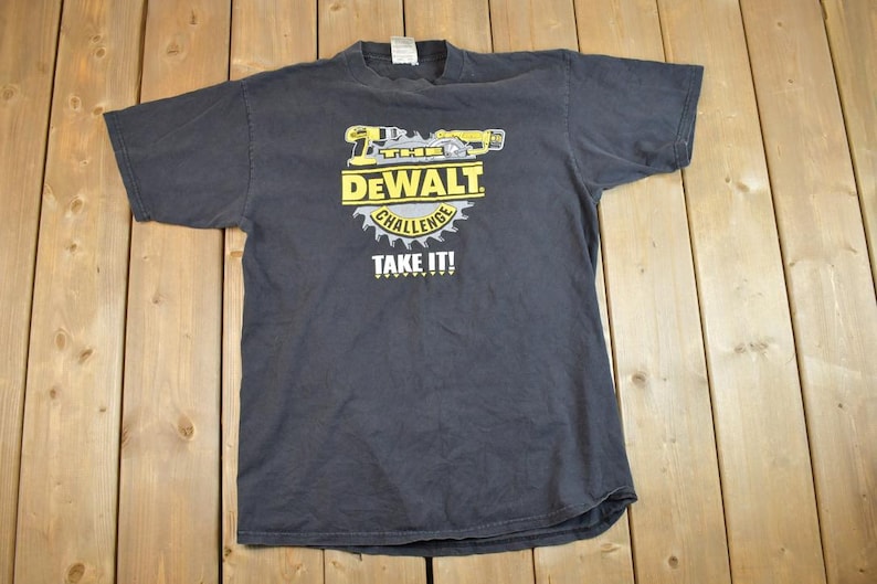 May include: A black t-shirt featuring a yellow and black Dewalt logo. The logo depicts a power tool and the text "Dewalt Challenge Take It!"