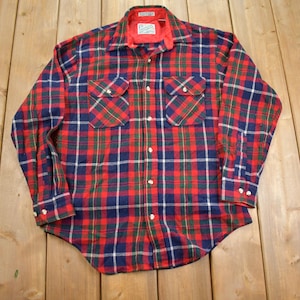 May include: A red, blue and green plaid flannel shirt with a button-down collar and long sleeves. The shirt has two chest pockets and a label that reads "Pacific Trail" on the left side of the collar.