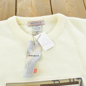May include: Cream-colored sweater with a crew neck and ribbed collar. The sweater has a tag that reads "AVIREX" and a rectangular patch at the bottom. The sweater is made of a soft, knitted material.