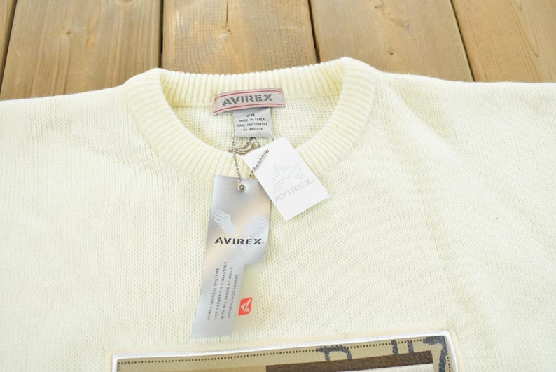 May include: Cream-colored sweater with a crew neck and ribbed collar. The sweater has a tag that reads "AVIREX" and a rectangular patch at the bottom. The sweater is made of a soft, knitted material.
