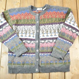 May include: Grey cardigan with a colourful landscape pattern. The button-down sweater features a design of trees, mountains, and floral elements in shades of red, green, blue, and pink. The sweater has a round neckline and long sleeves.