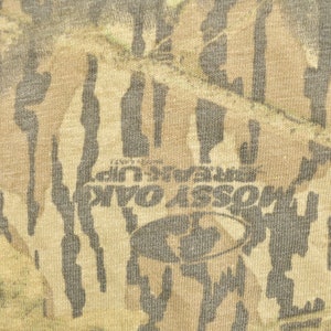 May include: Close-up of a fabric with a camouflage pattern in shades of brown, tan, and green. The text "MOSS OAK BREAK UP" is visible, along with the Mossy Oak logo. The fabric has a textured appearance.