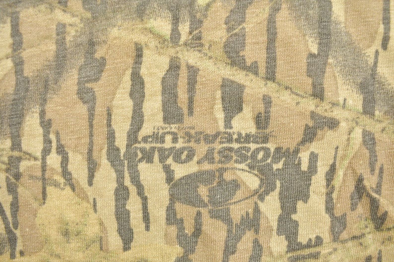 May include: Close-up of a fabric with a camouflage pattern in shades of brown, tan, and green. The text "MOSS OAK BREAK UP" is visible, along with the Mossy Oak logo. The fabric has a textured appearance.