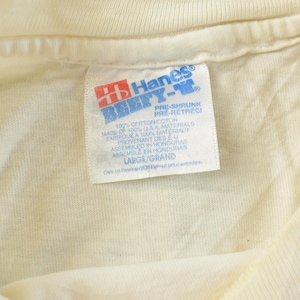 May include: Close-up of a cream-colored t-shirt collar with a Hanes BEEFY-T label. The label features red and blue text, with details about the shirt's material and size, including "LARGE/GRAND". The shirt is made of 100% cotton.