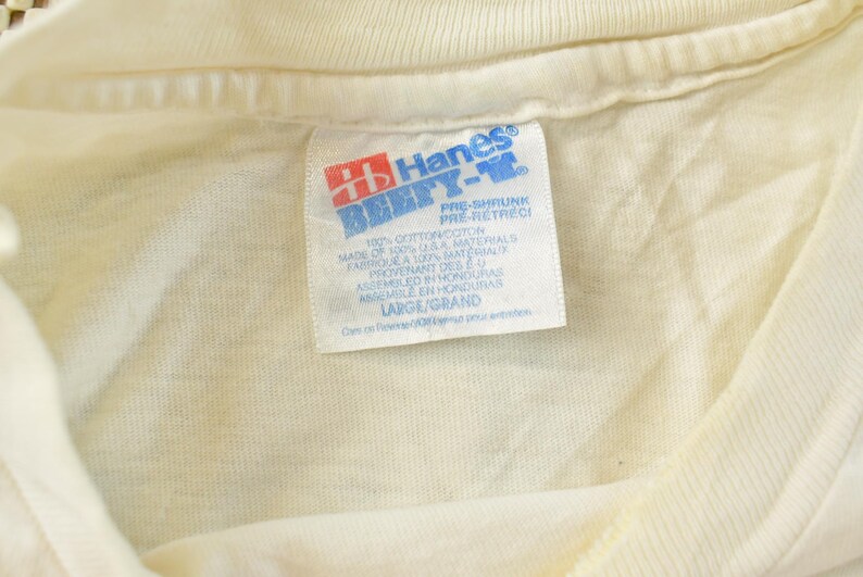 May include: Close-up of a cream-colored t-shirt collar with a Hanes BEEFY-T label. The label features red and blue text, with details about the shirt's material and size, including "LARGE/GRAND". The shirt is made of 100% cotton.