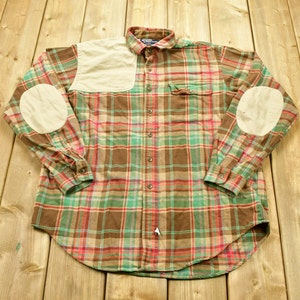 Könnte beinhalten: Ein braunes, grünes und rotes kariertes Flanellhemd mit Ellbogenpatches. Das Hemd hat einen Knopfkragenshirt und eine einzelne Brusttasche.