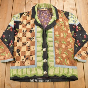 May include: A black, brown, green and blue patterned cardigan with a button closure. The cardigan has a chequered pattern, floral designs and a "Painted Pony" label.