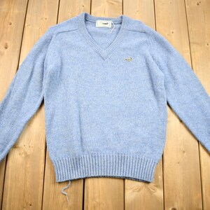 May include: A light blue v-neck sweater with long sleeves and a small green crocodile logo on the left chest.