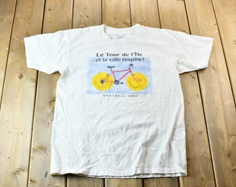 May include: White t-shirt with a graphic of a red bicycle with yellow wheels and the text "Le Tour de l'Ile...et la ville respire! Montreal 1987"