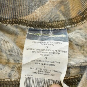 Puede incluir: Primer plano de una tela con estampado de camuflaje con una etiqueta adjunta. La etiqueta incluye texto en espa&ntilde;ol, franc&eacute;s e ingl&eacute;s, que detalla la composici&oacute;n de la tela y las instrucciones de cuidado. La tela es una mezcla de algod&oacute;n y poli&eacute;ster.