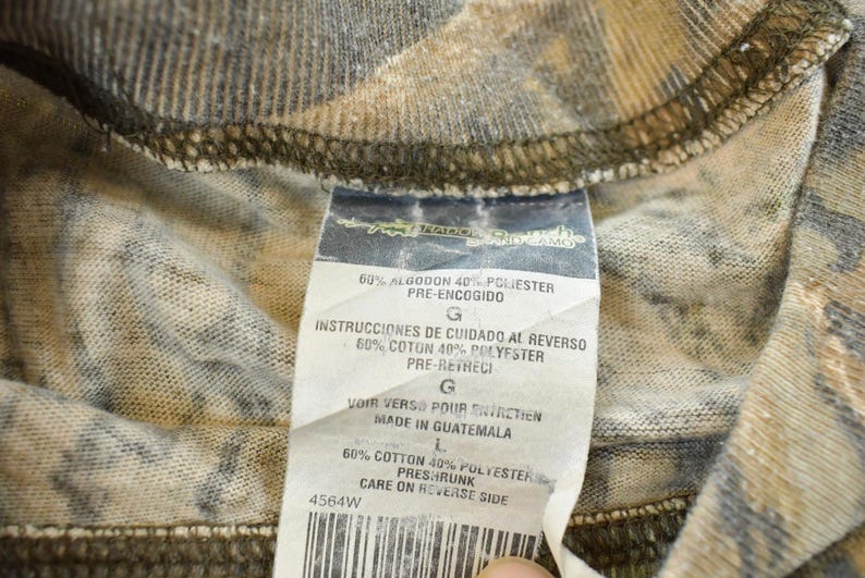 Puede incluir: Primer plano de una tela con estampado de camuflaje con una etiqueta adjunta. La etiqueta incluye texto en espa&ntilde;ol, franc&eacute;s e ingl&eacute;s, que detalla la composici&oacute;n de la tela y las instrucciones de cuidado. La tela es una mezcla de algod&oacute;n y poli&eacute;ster.