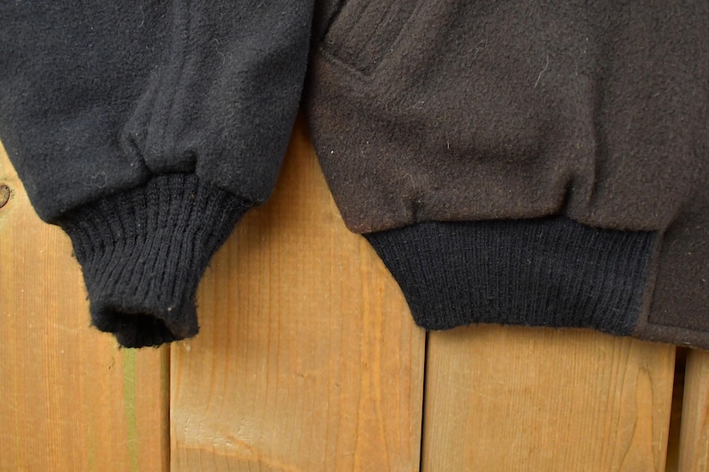 May include: Two jacket sleeves made of a thick, textured fabric. The left sleeve is black with a ribbed black knit cuff, while the right sleeve is dark brown with a matching ribbed black knit cuff. Both sleeves feature a visible seam detail.
