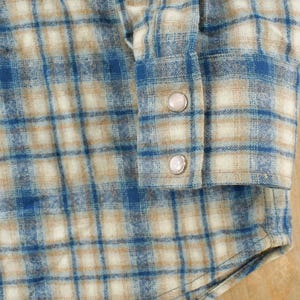 May include: Close-up of a blue, tan, and white plaid flannel shirt. The shirt features a buttoned cuff with two pearl-like buttons. The fabric has a soft, textured appearance, and the plaid pattern is clearly visible.