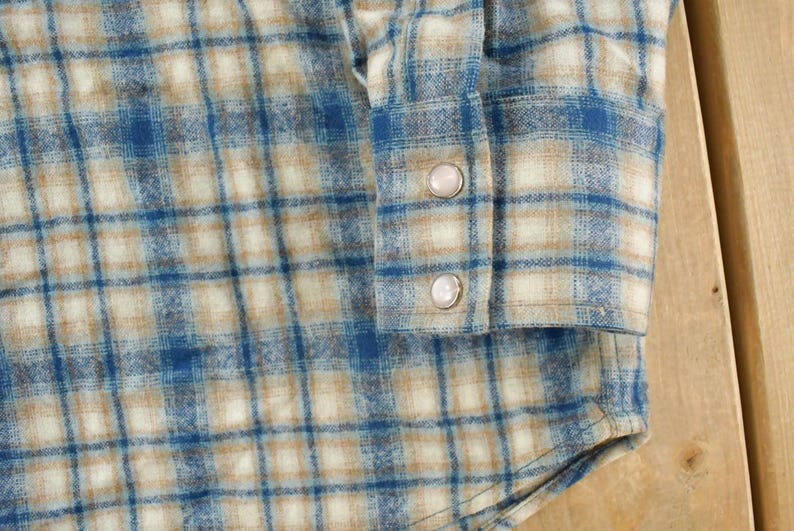 May include: Close-up of a blue, tan, and white plaid flannel shirt. The shirt features a buttoned cuff with two pearl-like buttons. The fabric has a soft, textured appearance, and the plaid pattern is clearly visible.
