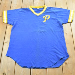 May include: Blue vintage baseball jersey with a yellow "P" on the chest. The jersey has a V-neck collar and sleeve cuffs with yellow and white stripes. The shirt is made of a soft material.