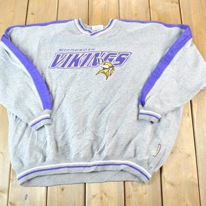 May include: A gray Minnesota Vikings sweatshirt with purple accents. The sweatshirt features the team's logo and text in purple and gold. The sleeves and neckline have purple stripes, and the cuffs have white and purple stripes.