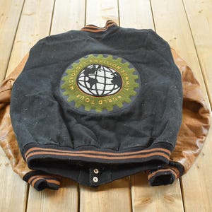 Könnte beinhalten: Schwarze und braune Vintage-Collegejacke mit Lederärmeln. Auf der Rückseite befindet sich ein Aufnäher mit einem Globus-Design und dem Text "Microsoft Envision Summit World Tour". Die Jacke hat eine Knopfleiste und gestreifte Verzierungen.