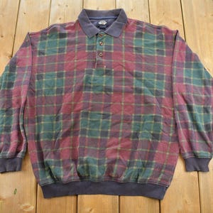 May include: A long-sleeve polo shirt featuring a plaid pattern in shades of maroon, dark green, and navy blue. The shirt has a solid navy blue collar, a three-button placket, and ribbed cuffs and hem, providing a structured and classic appearance.