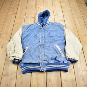 Puede incluir: Una chaqueta de béisbol de mezclilla azul con forro de sudadera con capucha. Las mangas son de color crema con rayas azules y blancas en los puños. La chaqueta tiene cierre a presión y dos bolsillos.